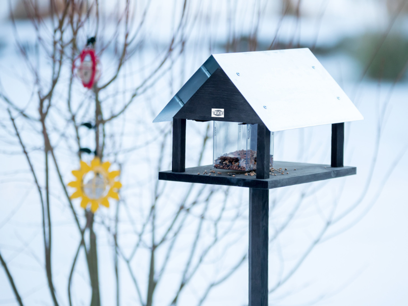 A black feeder with a metal roof for garden birds stands outside in the snow. In the background in the bare bushes hang two more feeders for tit dumplings.