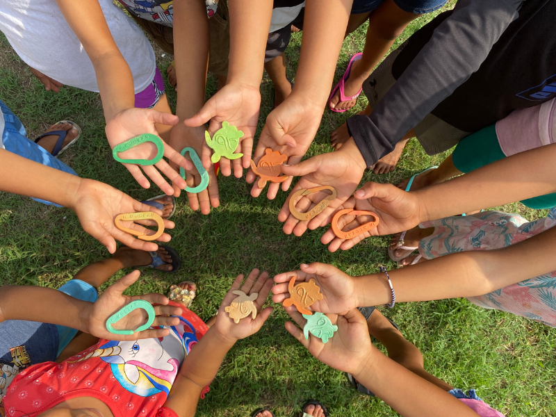 Children hold small plastic rakes and plastic turtles in a circle in the palms of their hands.