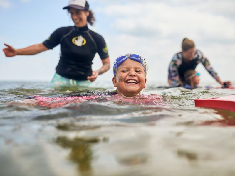 A child is swimming in the sea and laughing heartily. His swimming instructor is standing behind him.