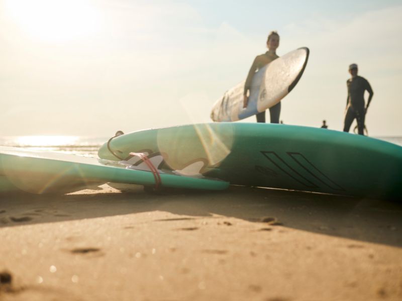 Two surfboards lie on the beach. In the background, a surfer stands with a board under his arm.