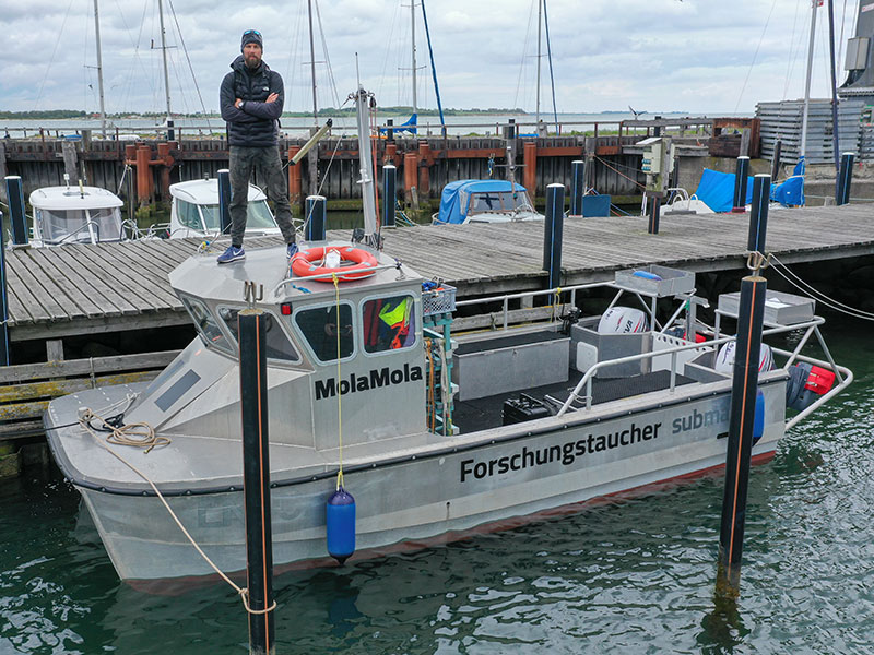 Ein Mann steht auf einem Boot namens Forschungstaucher, welches am Bootssteg liegt. 