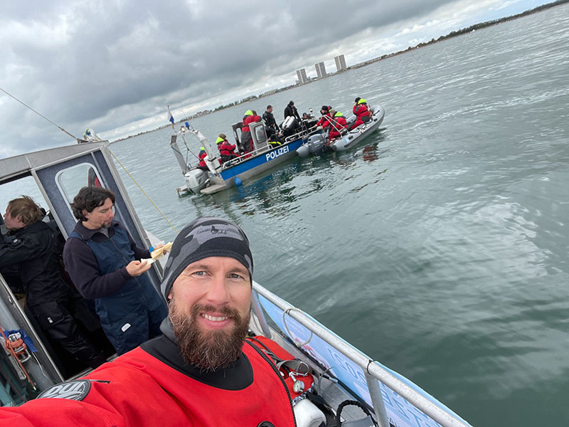 A man in a red jacket takes a selfie on a ship. In the background, an inflatable boat with several people.	