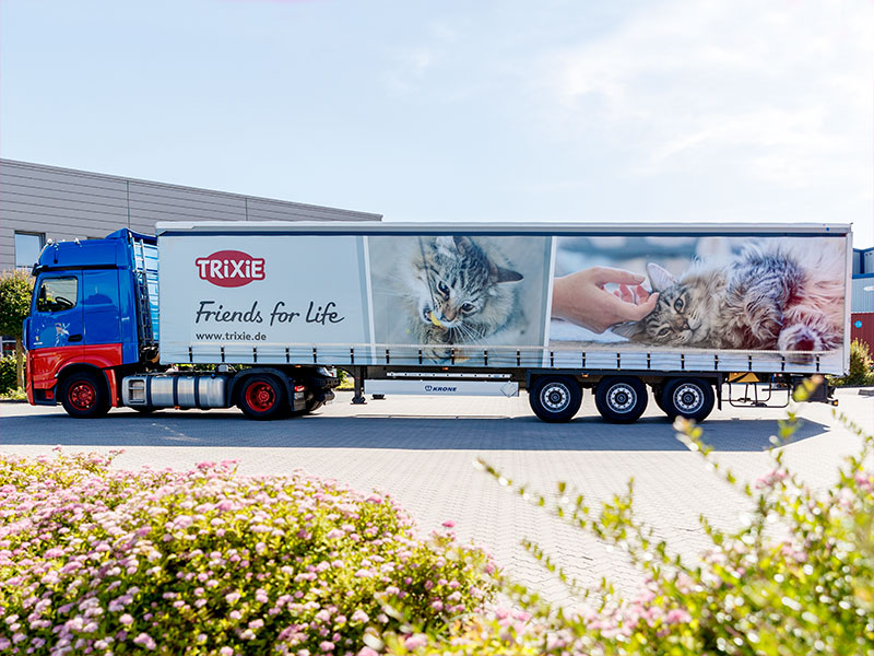 Vrachtwagen met TRIXIE-logo en de slogan "Friends for Life" aan de zijkant. Afbeeldingen van huisdieren sieren de trailer. De vrachtwagen staat geparkeerd op een parkeerplaats.